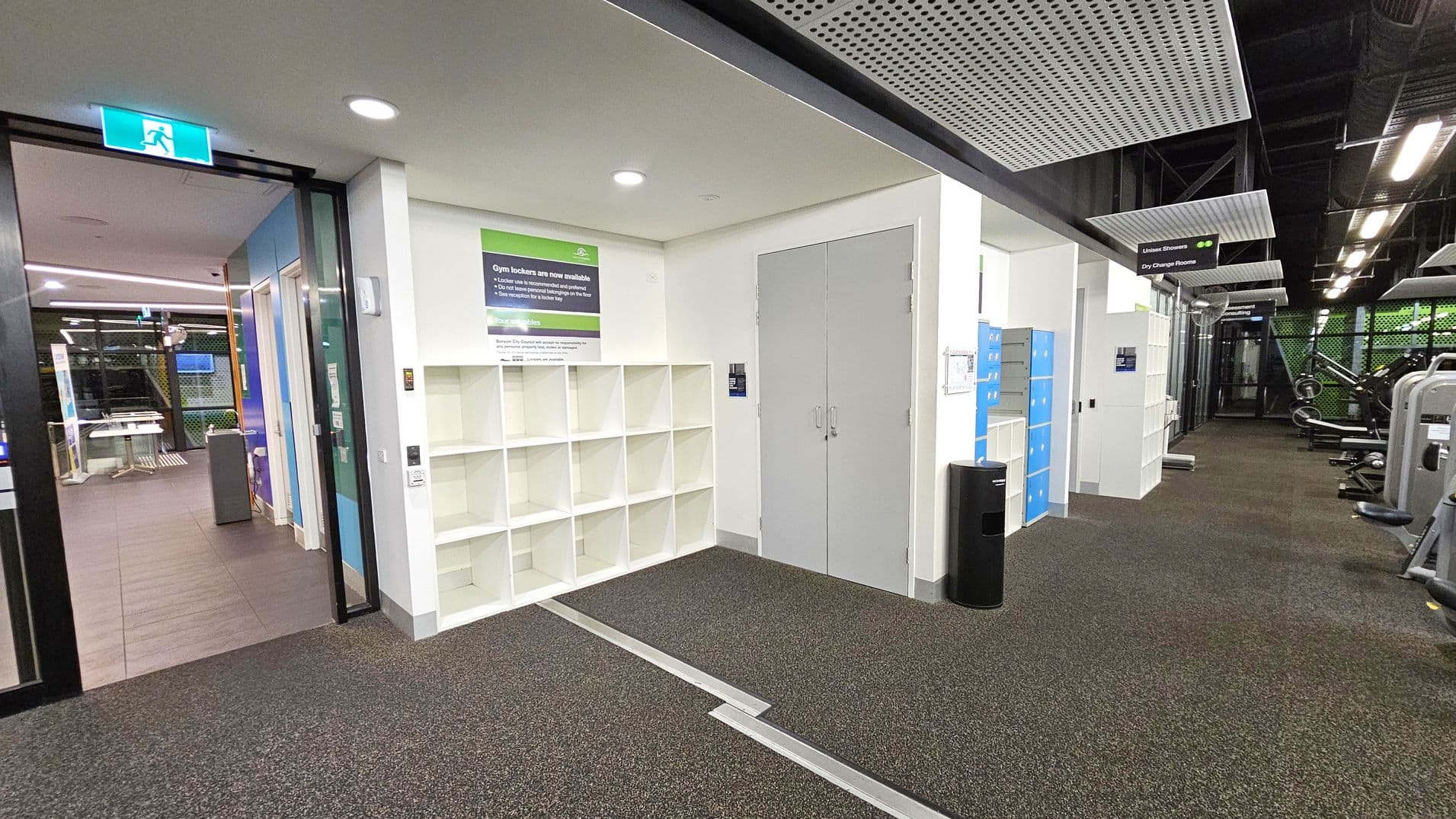 On the left, there's a glass door with an illuminated exit sign above it, leading to a room with tables and chairs. In the centre, white cubby shelves are mounted on the wall beneath a sign about gym lockers. To the right, blue lockers and gym equipment are visible. The floor is carpeted in a speckled pattern, and the ceiling features perforated panels with exposed ductwork.