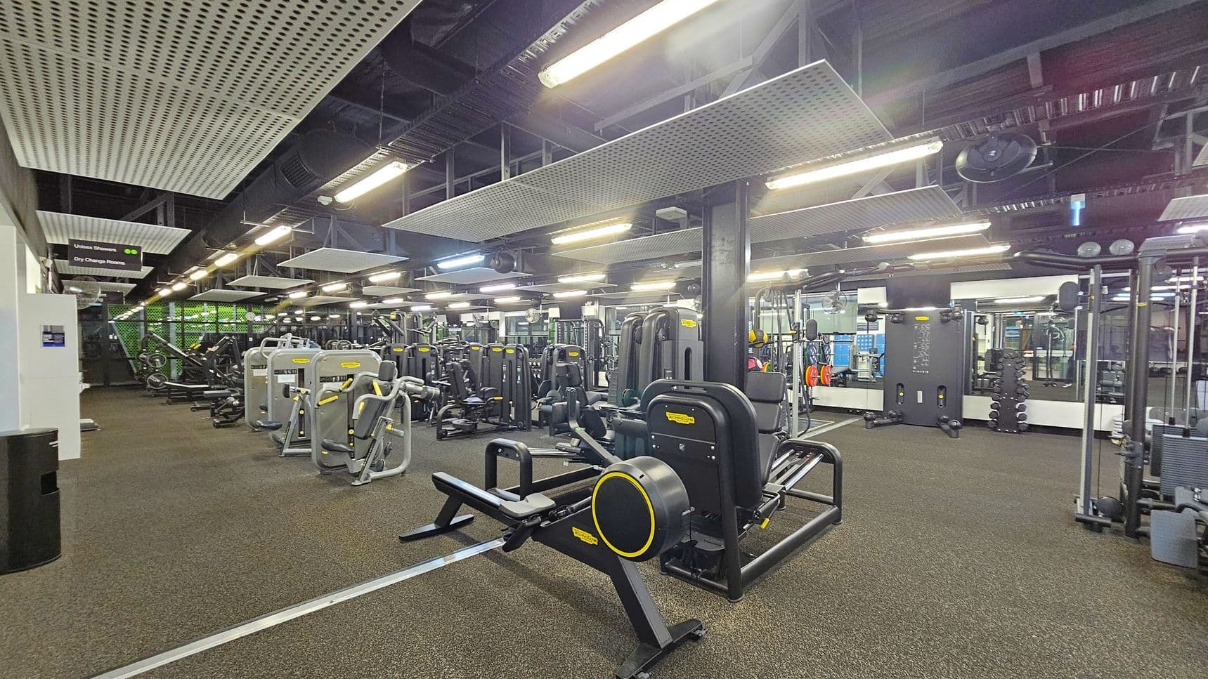 A spacious gym interior with various exercise machines arranged in rows. On the left, there are signs for union showers and dry change rooms. The floor is covered with a speckled rubber mat, and the ceiling features large, rectangular acoustic panels and fluorescent lights. In the centre, there are multiple strength training machines, and on the right, a wall with mirrors and additional equipment, including weights and resistance machines.