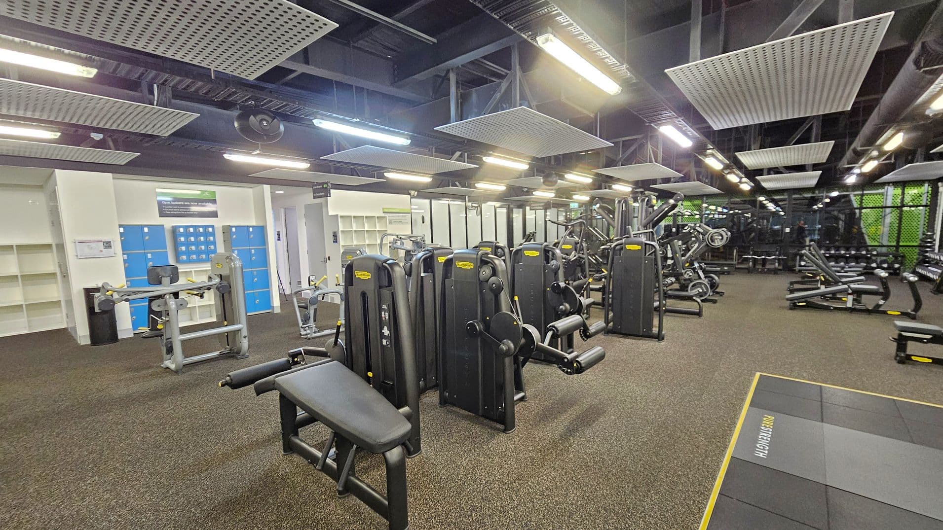 A spacious gym with various exercise machines. On the left, there are blue lockers and shelves against a light-coloured wall. In the centre, multiple black and grey workout machines are arranged in rows. To the right, more equipment, including benches and weights, is visible. The floor is covered with brown carpet tiles, and the ceiling features exposed beams and rectangular light fixtures.