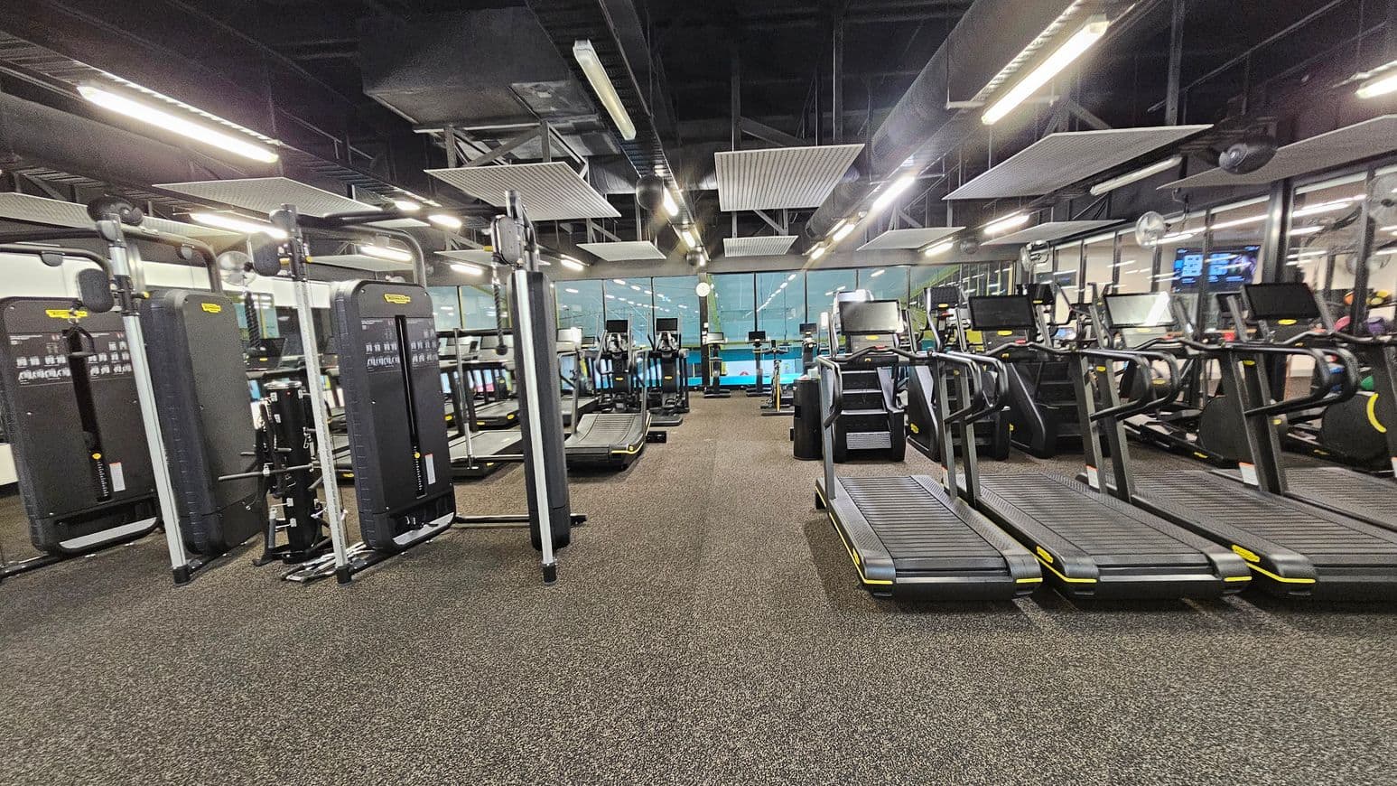 A gym with a variety of exercise equipment. On the left, there are several weight machines with black panels and metal frames. In the centre, there are multiple treadmills and elliptical machines lined up, facing large windows. On the right, more treadmills are visible. The floor is covered with a speckled brown and green carpet. The ceiling is high with exposed ductwork and rectangular light fixtures.