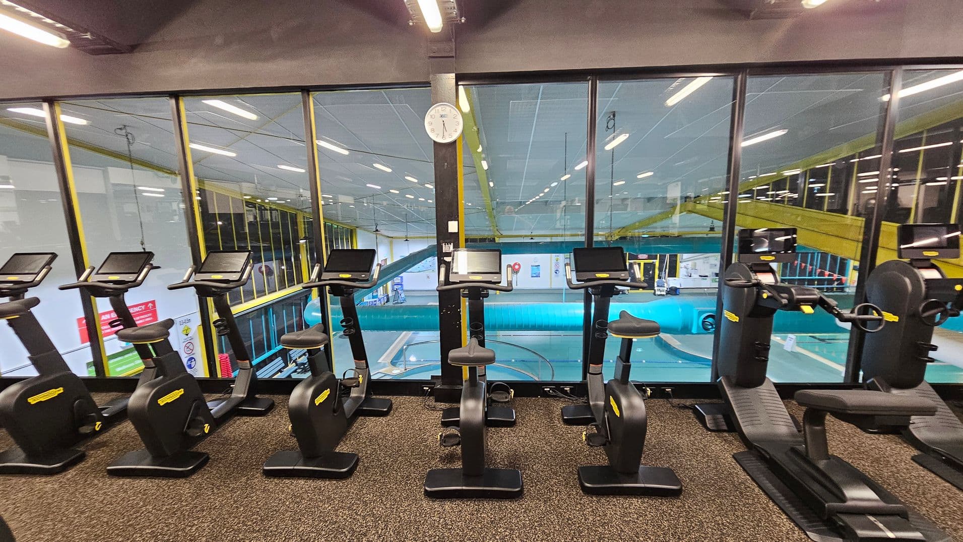 A row of stationary exercise bikes and recumbent bikes with digital screens is positioned on a speckled carpeted floor. Behind them, large glass windows reveal an indoor swimming pool area with a blue water slide and yellow railings. A white clock is mounted on a black pillar in the centre of the window. The ceiling has fluorescent lights, and the overall setting is a gym overlooking a pool area.