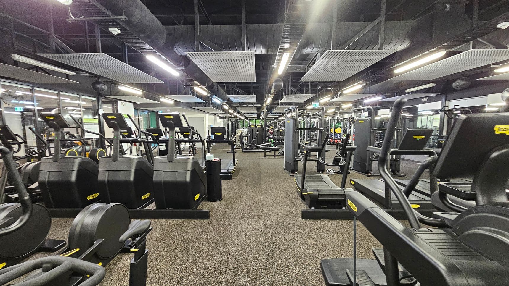 A spacious gym interior with rows of black exercise machines, including treadmills and ellipticals, on both sides. The floor is covered with brown carpet tiles. The ceiling is high with exposed ductwork and rectangular acoustic panels. Bright fluorescent lights illuminate the space. In the background, there are additional fitness equipment and weightlifting areas. Large mirrors line the walls, reflecting the equipment and enhancing the sense of space.