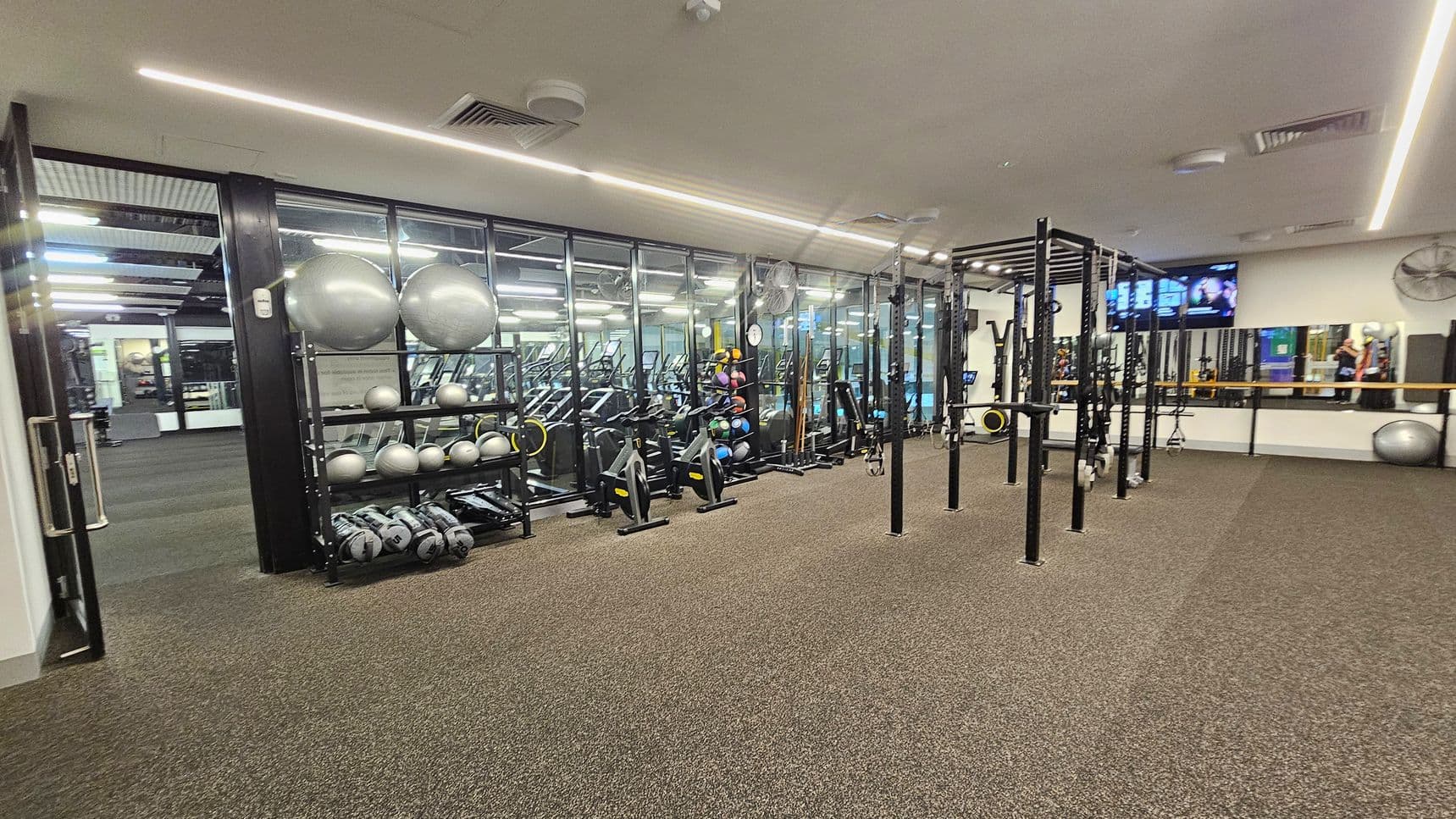 A gym with a brown carpeted floor and a ceiling with recessed lighting. On the left, there is a rack with silver exercise balls and medicine balls. In the middle, there are stationary bikes and a large metal frame for exercises. On the right, a wall-mounted TV and a mirror reflect the gym equipment. The background features glass walls separating this area from another section of the gym.