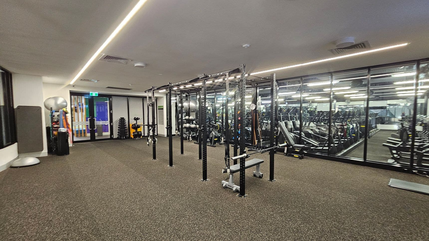 A gym with a brown carpeted floor and a white ceiling featuring linear lights. On the left, there are exercise balls, mats, and a doorway. In the centre, a large metal frame structure with various gym equipment like benches and straps. On the right, a mirrored wall reflects rows of treadmills and other cardio machines.