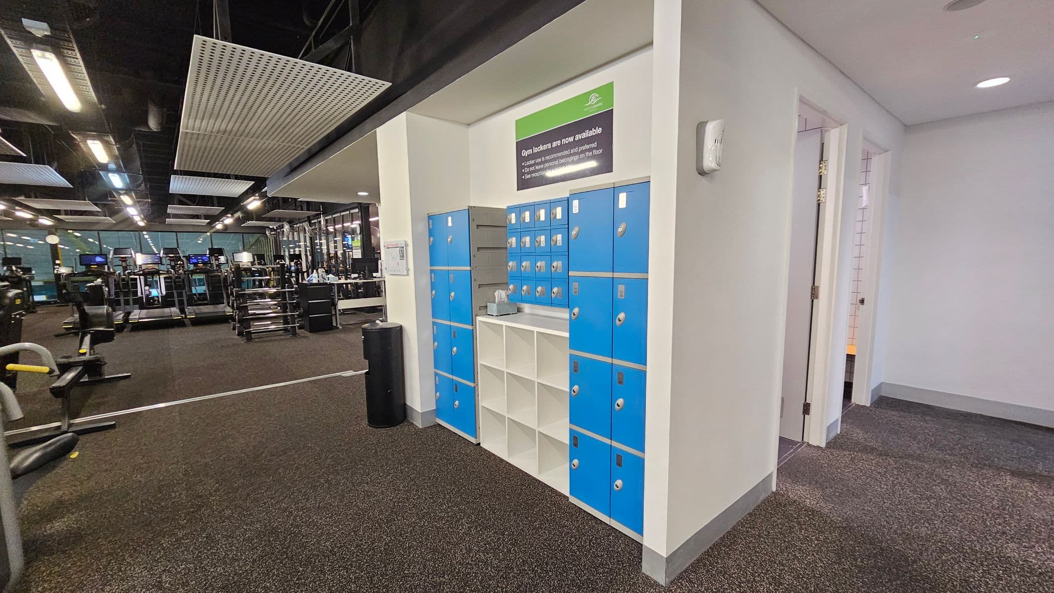 On the left, there is a gym area with various exercise machines, including treadmills and weightlifting equipment. The floor is covered with dark carpeting. In the middle, there is a section with blue lockers and open white cubbies. A sign above the lockers indicates their availability. On the right, there is a corridor with open doors leading to other rooms. The ceiling has a modern design with rectangular light fixtures and panels.