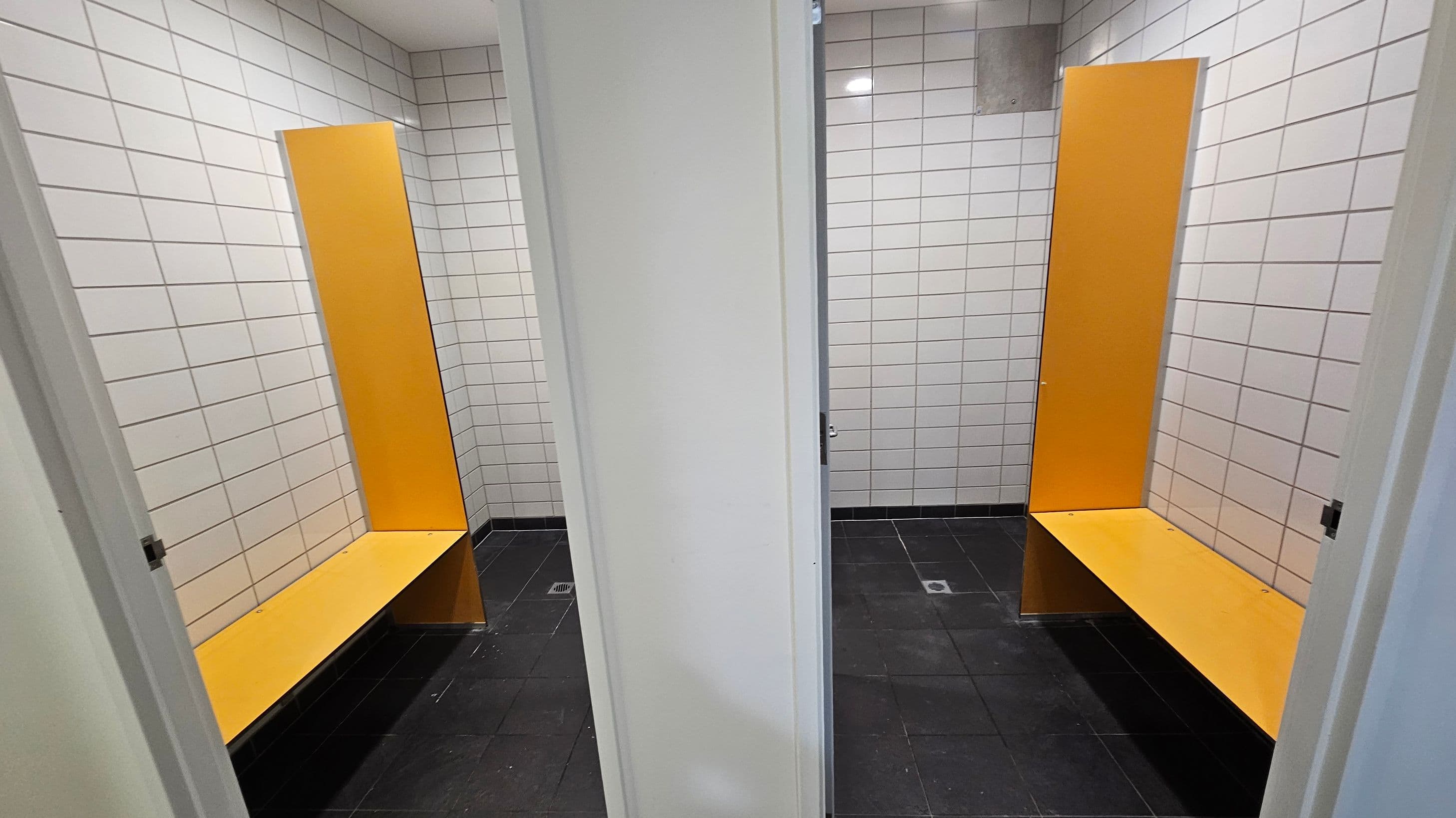 Two changing cubicles are visible, separated by a white partition. Each cubicle has white tiled walls and a black tiled floor. Inside each cubicle, there is a bright orange bench with a matching vertical panel behind it. The ceiling is plain white with recessed lighting.