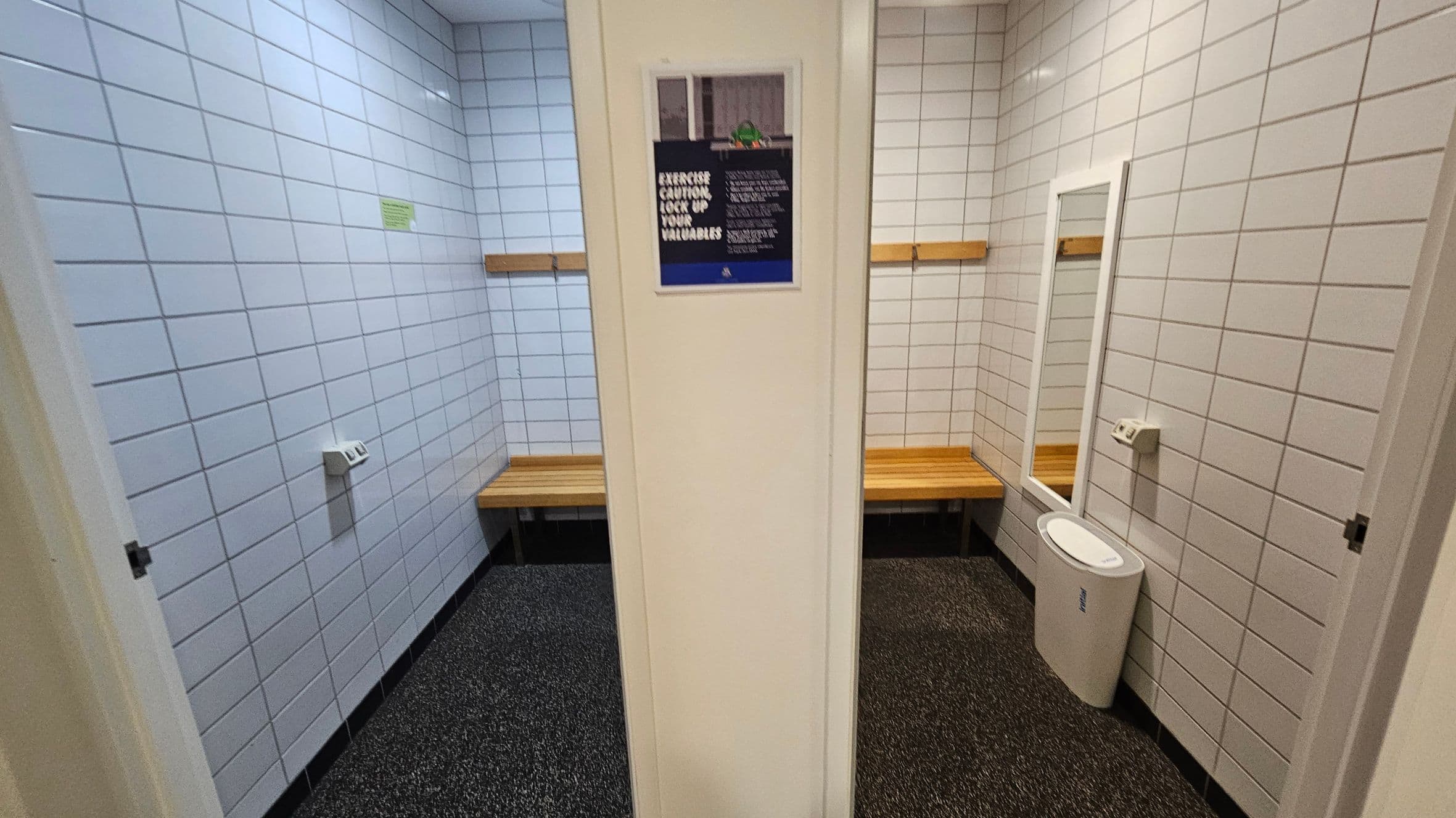 A changing room with white tiled walls and a speckled floor. On the left, there is a wooden bench beneath a coat hook. In the centre, a wall with a sign. On the right, another wooden bench, a full-length mirror, and a white bin. The ceiling is plain with recessed lighting.