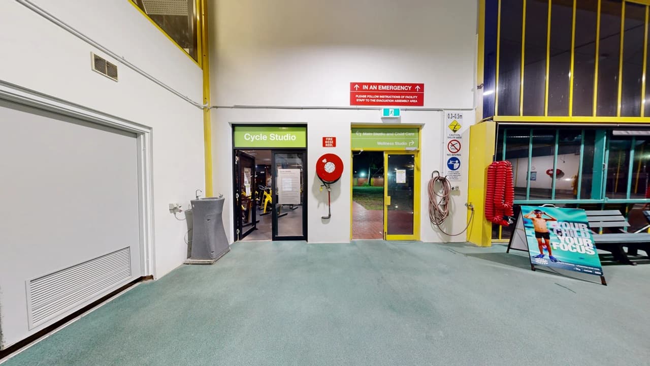 A hallway with a green carpeted floor. On the left, there's a water fountain and a door labelled "Cycle Studio" with exercise equipment visible inside. In the centre, a yellow-framed glass door leads outside, with a fire hose reel mounted on the wall. On the right, a coiled hose and a sign with safety instructions are visible. A poster stand with an image of a person is near a bench. The walls are white with yellow accents, and the ceiling is not visible.