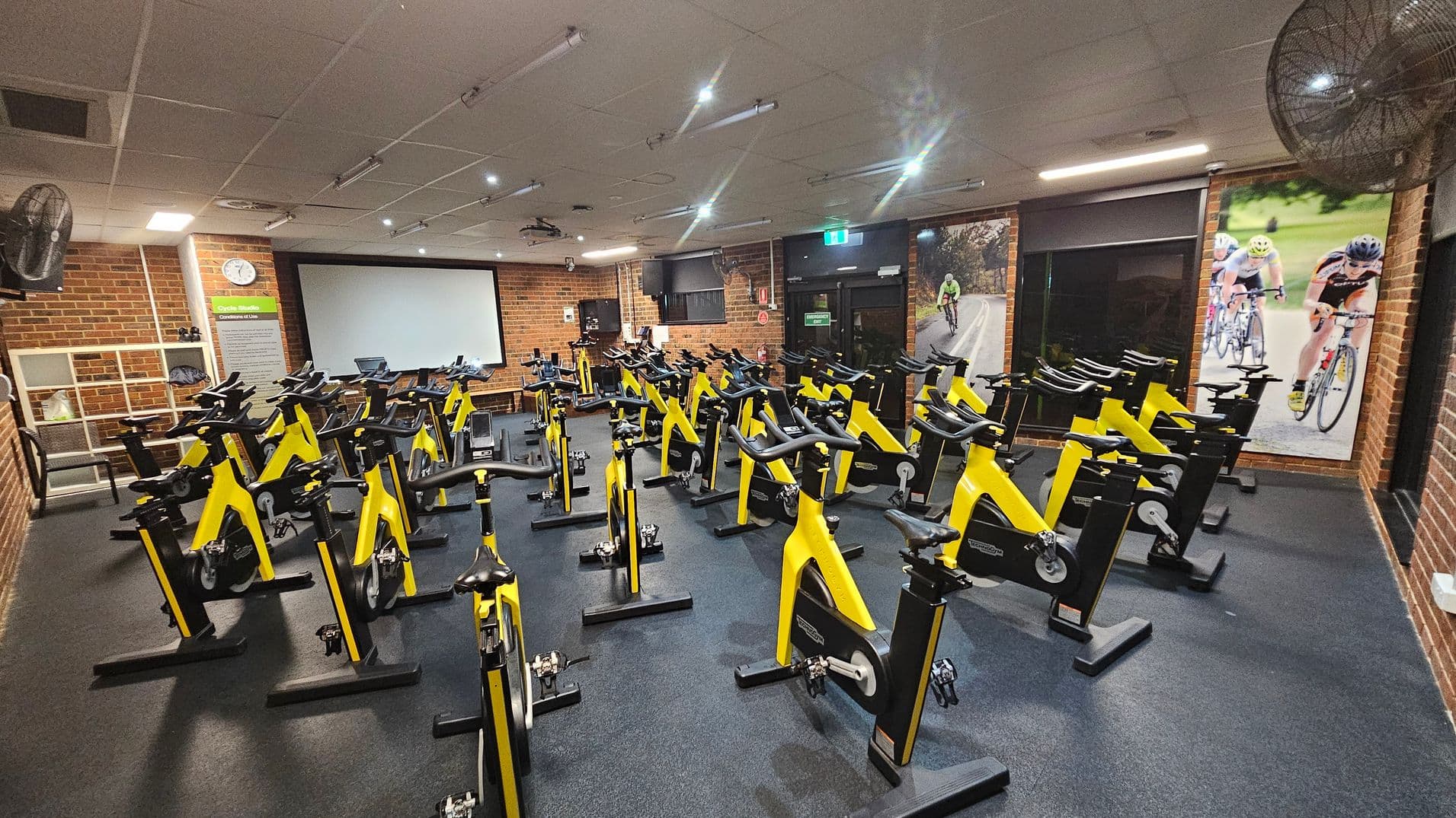 A cycling studio with numerous yellow stationary bikes arranged in rows on a dark floor. The walls are brick, with large posters of cyclists on the right. At the back, there's a large screen and a clock on the left wall. The ceiling is fitted with lights and fans.