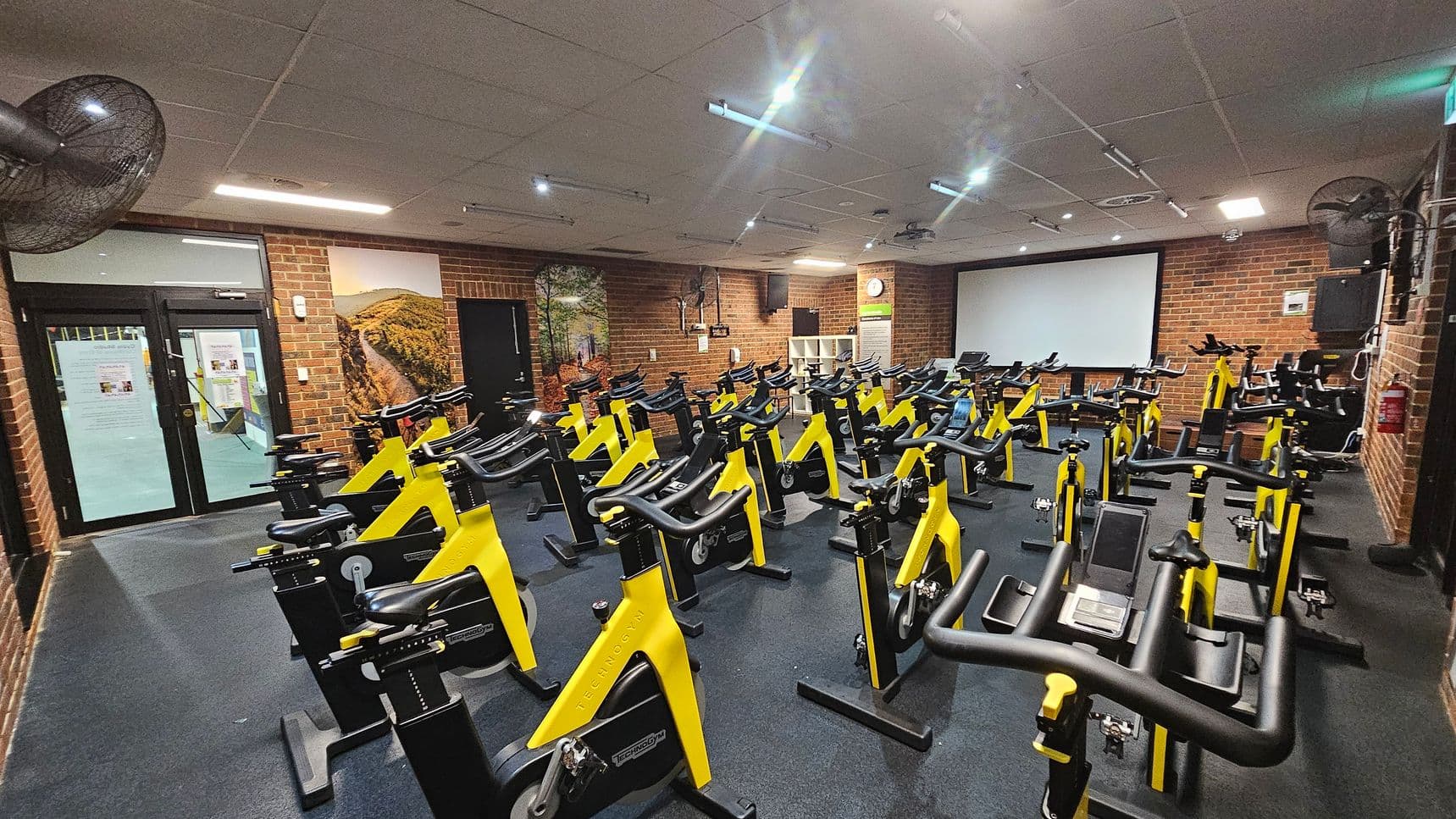 A gym room with numerous yellow and black stationary bikes arranged in rows on a dark floor. The walls are brick, with a large screen at the front. On the left, there are glass doors with notices and a wall mural depicting a landscape. Ceiling fans and lights are visible above.