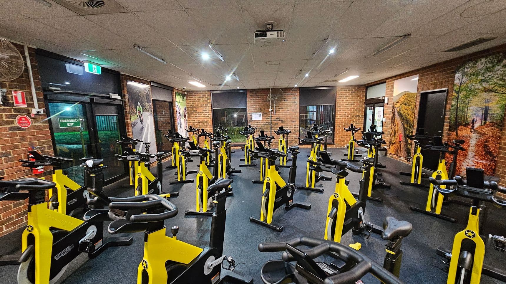 A room with numerous yellow stationary bikes arranged in rows on a dark floor. The walls are brick, featuring large cycling-themed murals. On the left, there's an emergency exit door with a green sign above it. The ceiling is fitted with fluorescent lights and a projector. The room is well-lit, and there are windows with blinds on the far wall.