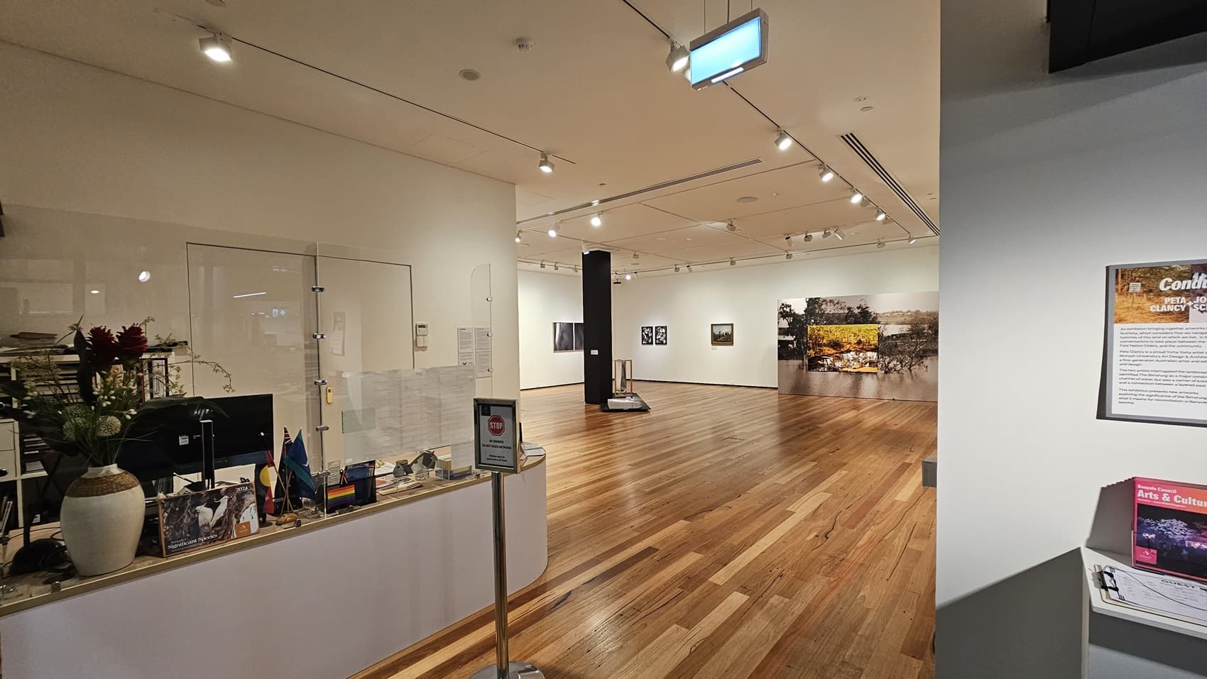 On the left, there is a white reception desk with a clear partition, flowers in a vase, and various brochures and items. In the middle, there is an open space with wooden flooring, leading to an exhibition area with artworks on the walls. In the background, there are more artworks and a display stand. On the right, there is a white wall with a framed information board and a small shelf holding brochures. The ceiling is white with recessed lighting and track lights.