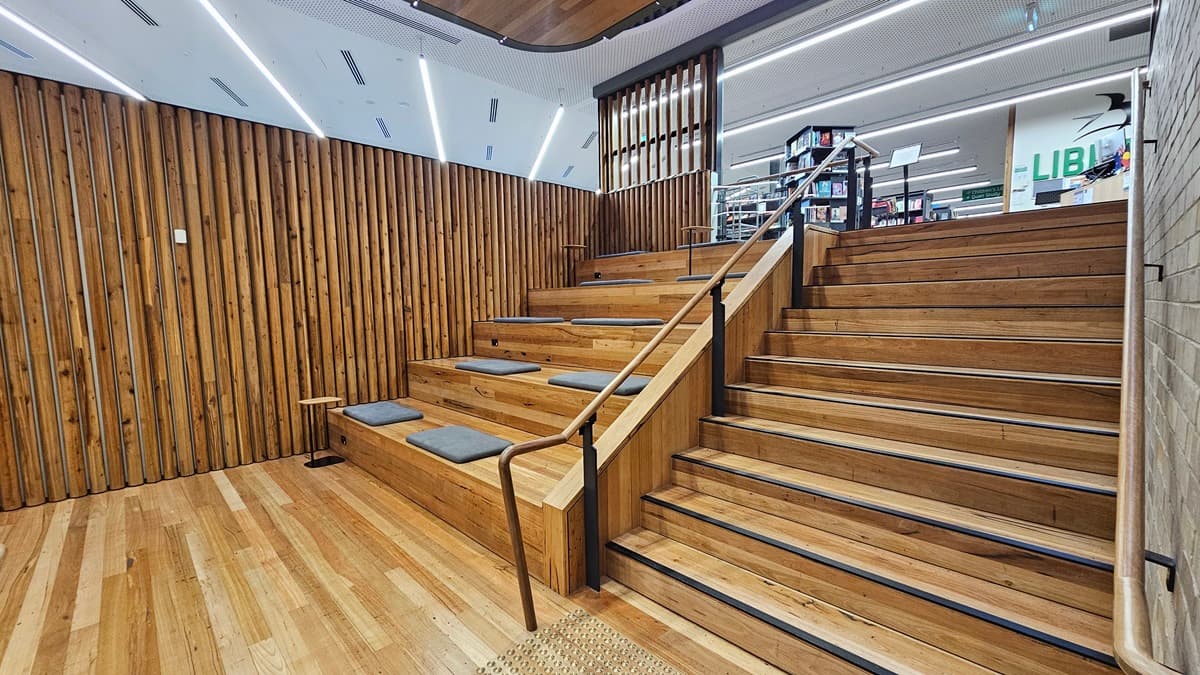 On the left, there is a lift with a "Staff Only" sign next to it, a grey column, and a long table with black chairs near large windows. In the middle, there is a staircase with wooden steps and rainbow-coloured stripes, leading to an upper level with a glass railing. On the right, there is a white wall with a door and a sign for "Workspaces." The floor is covered in grey tiles, and the ceiling is white with a perforated design and wooden panels with recessed lighting.