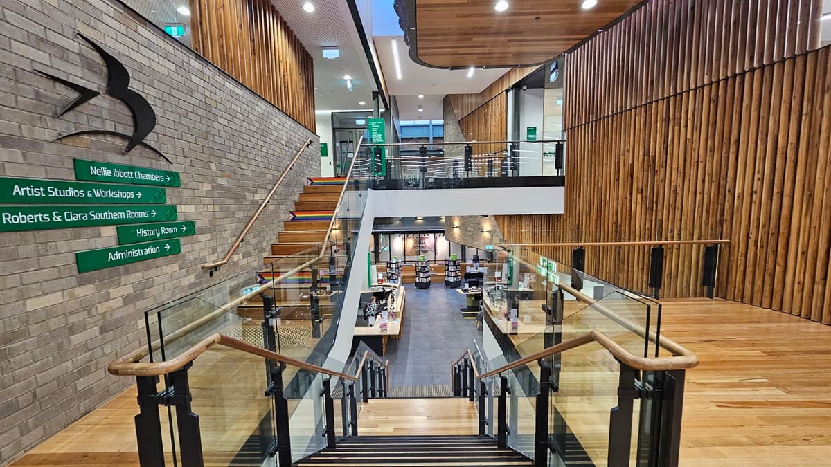 On the left, there is a brick wall with green directional signs and a black bird sculpture. In the middle, there is a staircase with wooden steps and glass railings leading down to a lower level with bookshelves and a reception desk. On the right, there is a wooden wall with vertical slats and a glass railing on the upper level. The floor is made of light-coloured wood, and the ceiling has white panels with recessed lighting and wooden sections.