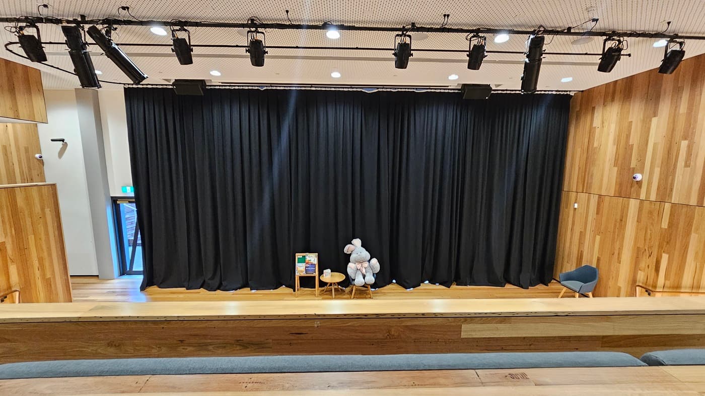 On the left, there is a wooden wall with vertical panels and a small exit door with a green emergency exit sign above it. On the right, another wooden wall with vertical panels extends to the ceiling. In the middle, there is a stage with black curtains, a small wooden table, a chair, and a large stuffed animal. The foreground features tiered wooden seating steps with grey cushions. The floor is wooden, and the ceiling is white with a perforated design and black stage lighting fixtures.