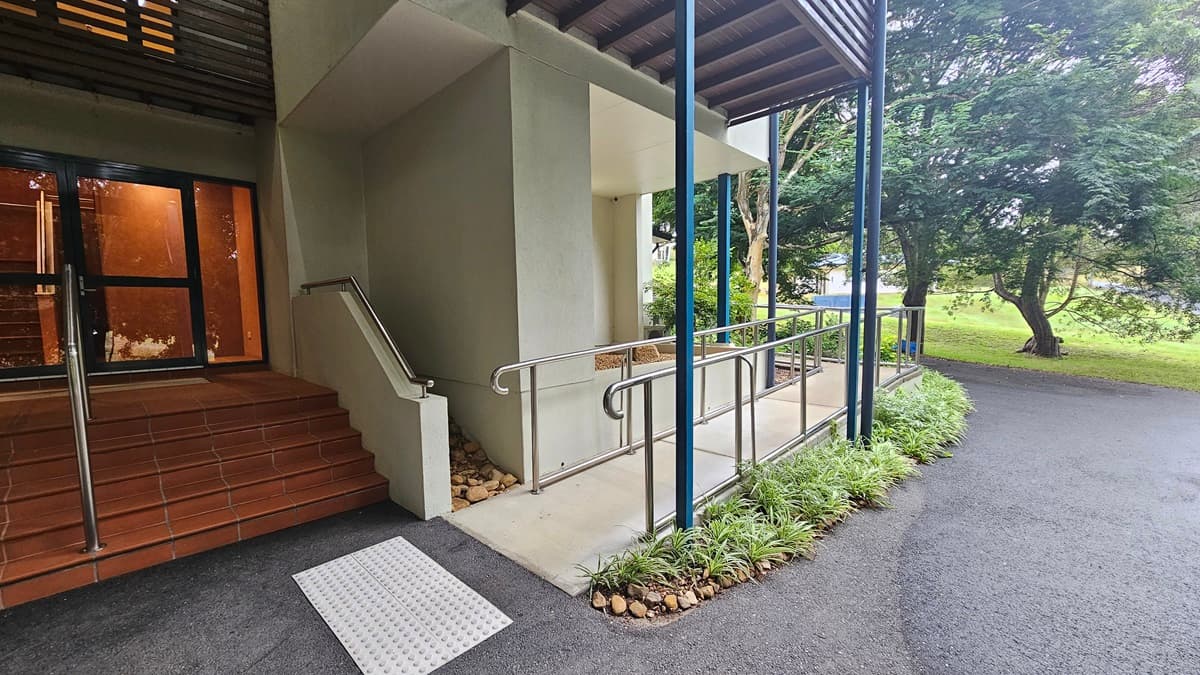On the left, there is a set of brown tiled steps leading to a glass door. A metal handrail runs alongside the steps. In the centre, a ramp with metal railings provides an accessible path, bordered by small plants and rocks. On the right, a paved path curves away, with trees and greenery in the background. The ceiling above the ramp is supported by blue metal beams.