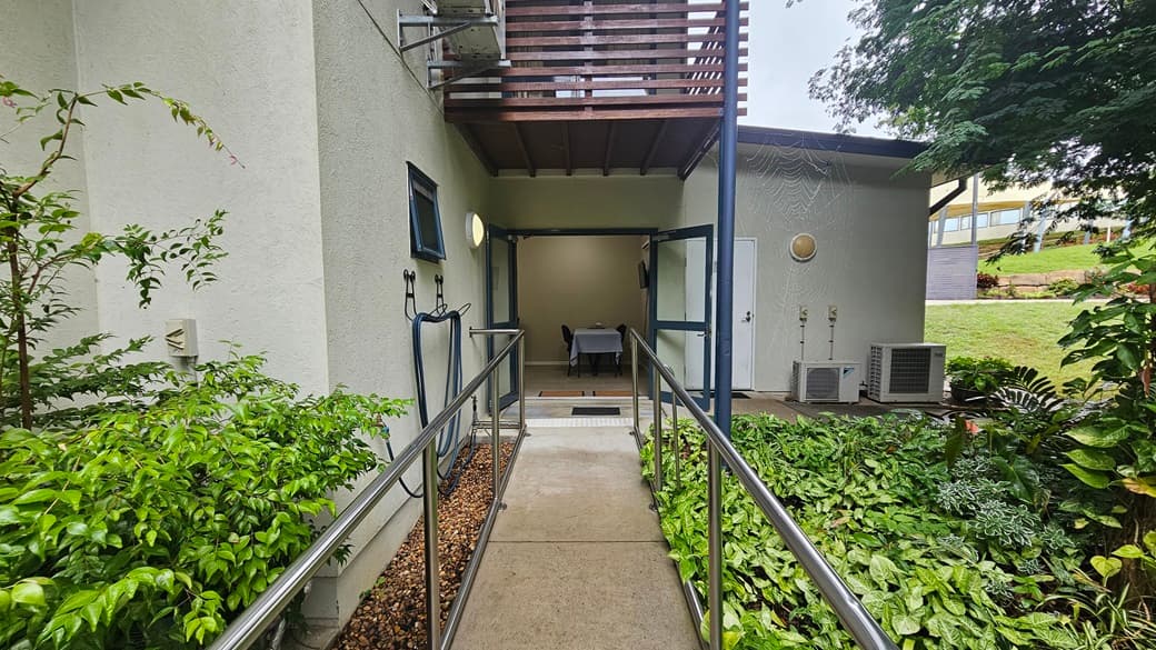 A narrow concrete path with metal railings leads to a building entrance. On the left, there are lush green plants and a small window on the building wall. On the right, more greenery is visible, along with air conditioning units and a door. Above the entrance, there is a wooden balcony. The background shows a grassy area with trees and a glimpse of another building.