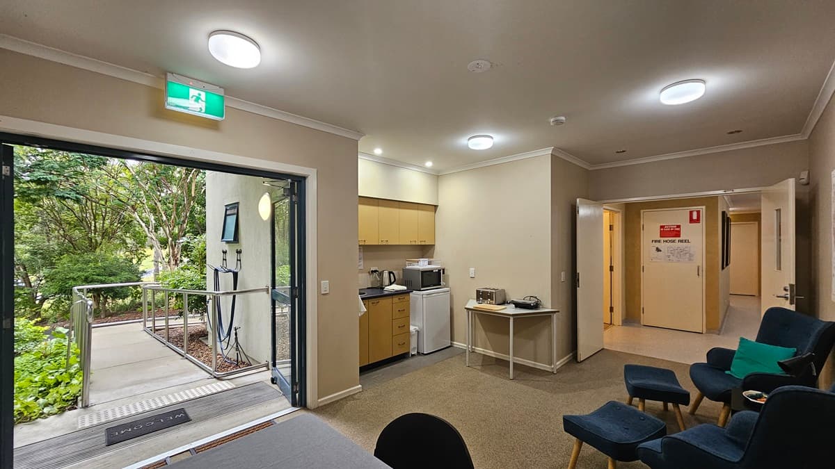 A room with beige walls and a carpeted floor. On the left, a glass door opens to a garden with trees and a ramp. In the middle, a kitchenette with wooden cabinets, a microwave, kettle, and mini-fridge. On the right, a hallway with open doors and a red emergency sign. Several dark blue chairs with teal cushions are arranged in the foreground. The ceiling has recessed lighting and a glowing exit sign above the door.