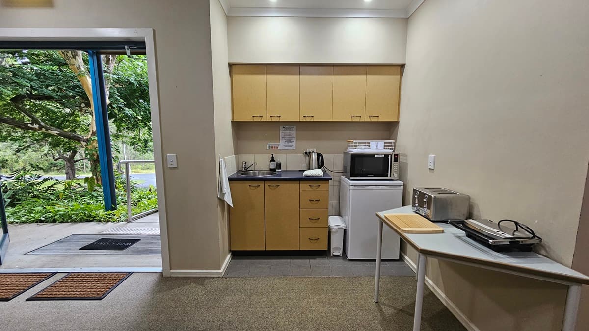 A small kitchen area with light wood cabinets and a black countertop. On the counter are a kettle and a microwave. Below is a mini fridge. To the right, a table holds a toaster and a sandwich press. The floor is carpeted, and the ceiling has recessed lighting. On the left, an open door leads outside, revealing lush greenery and a pathway.