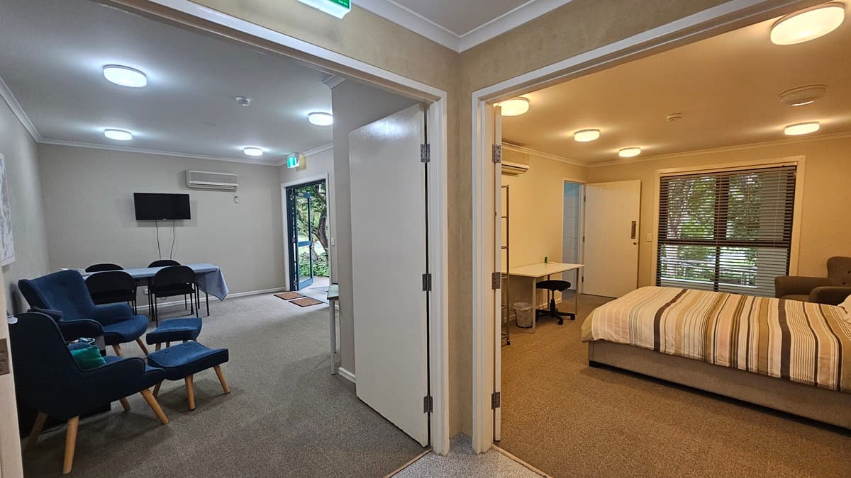 A view of two rooms from a hallway. On the left, a lounge area with blue chairs, a small table, a wall-mounted TV, and a glass door leading outside. On the right, a bedroom with a striped bed, a desk with a chair, and a window with blinds. Both rooms have beige walls and carpeted floors. Ceiling lights are visible in both spaces.