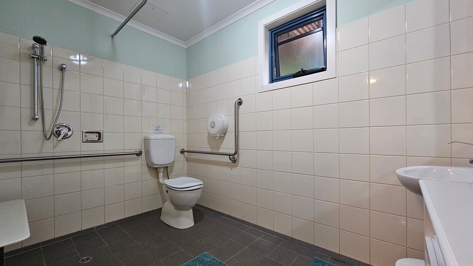 A bathroom with light-coloured tiled walls and a dark tiled floor. On the left, there is a shower with a handheld showerhead and a horizontal grab bar. In the centre, a toilet with a vertical grab bar beside it. Above the toilet, a small window with a blue frame. On the right, a white sink with a tap. The ceiling is white with a simple design.