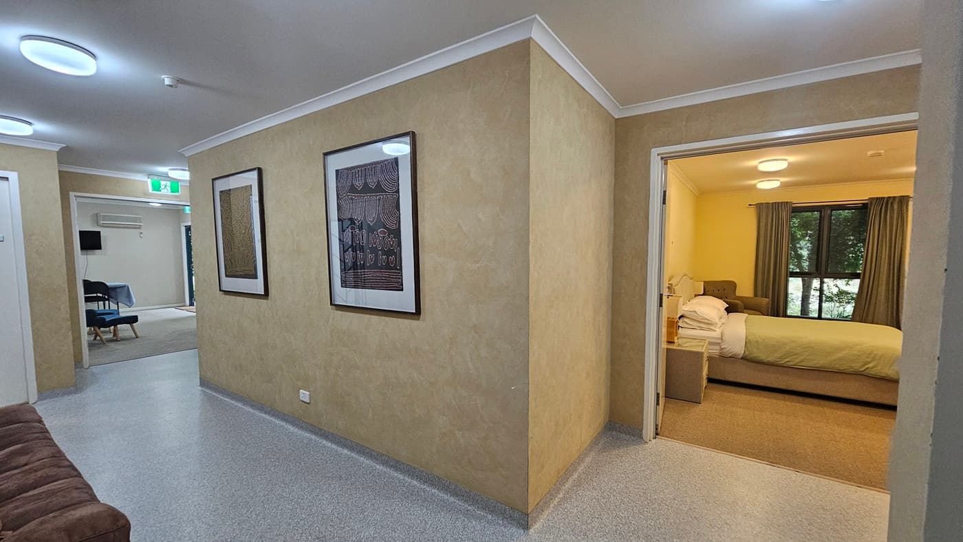 A hallway with light-coloured walls and a speckled floor. On the left wall, two framed artworks are displayed. To the right, an open doorway leads to a bedroom with a bed, bedside table, and a window with curtains. In the background, another room is visible with chairs and a table, and an exit sign above a door. The ceiling has recessed circular lights.
