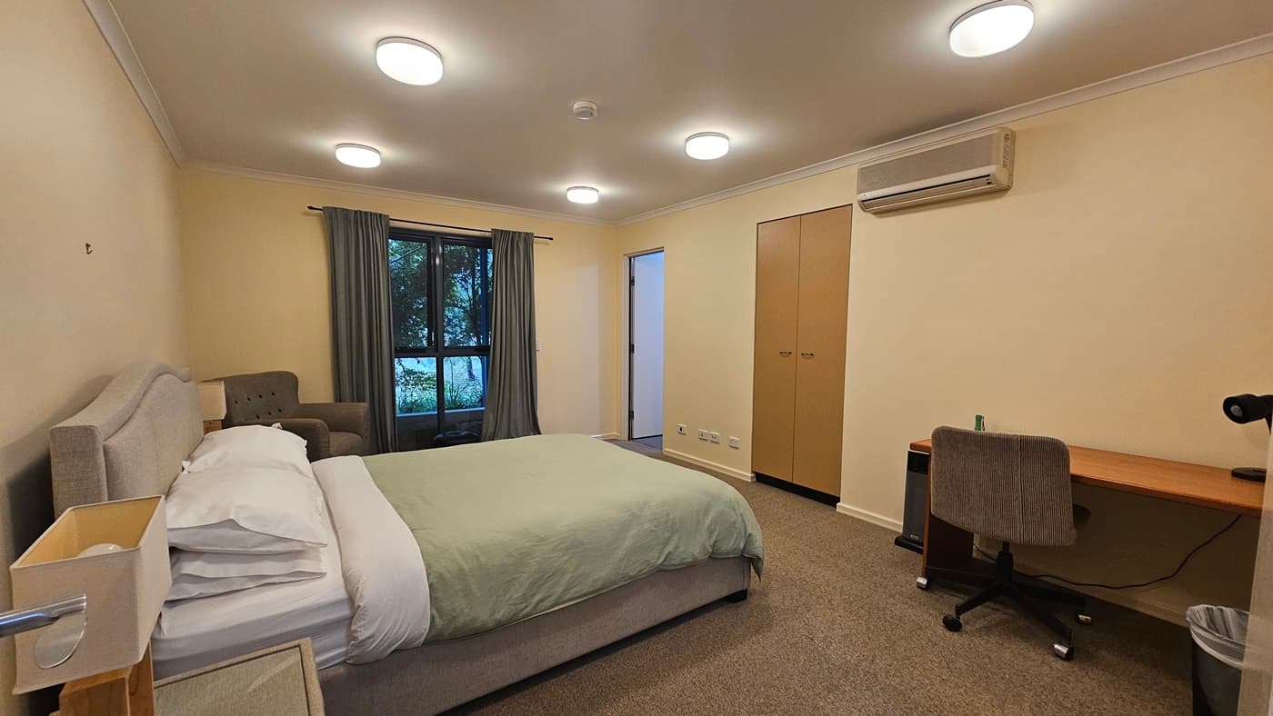 A bedroom with a bed on the left, covered with a green duvet and white pillows. Next to the bed is a grey armchair. A window with green curtains is in the background. On the right, there's a wooden desk with a grey office chair and a small bin underneath. Above the desk is an air conditioning unit. A wardrobe with two doors is next to the desk. The ceiling has several round lights, and the floor is carpeted.