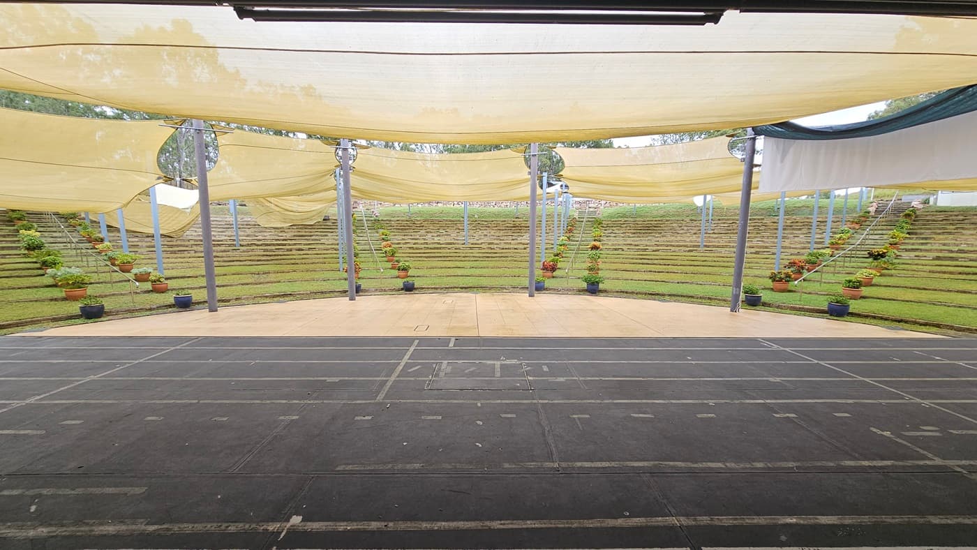An outdoor amphitheatre with tiered grass seating in the background, arranged in a semi-circle. The foreground features a dark, paved stage area with visible lines. Overhead, beige fabric canopies are stretched across metal poles, providing shade. Potted plants with flowers are placed along the steps and near the poles, adding greenery to the scene.