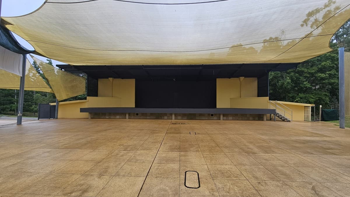 An outdoor stage with a large, flat, open area in front, covered by a series of beige fabric canopies. The stage is elevated and painted in a light yellow colour, with a black backdrop. There are steps on the right side leading up to the stage. The ground is paved with large, square tiles. Trees and greenery are visible in the background on the right side.
