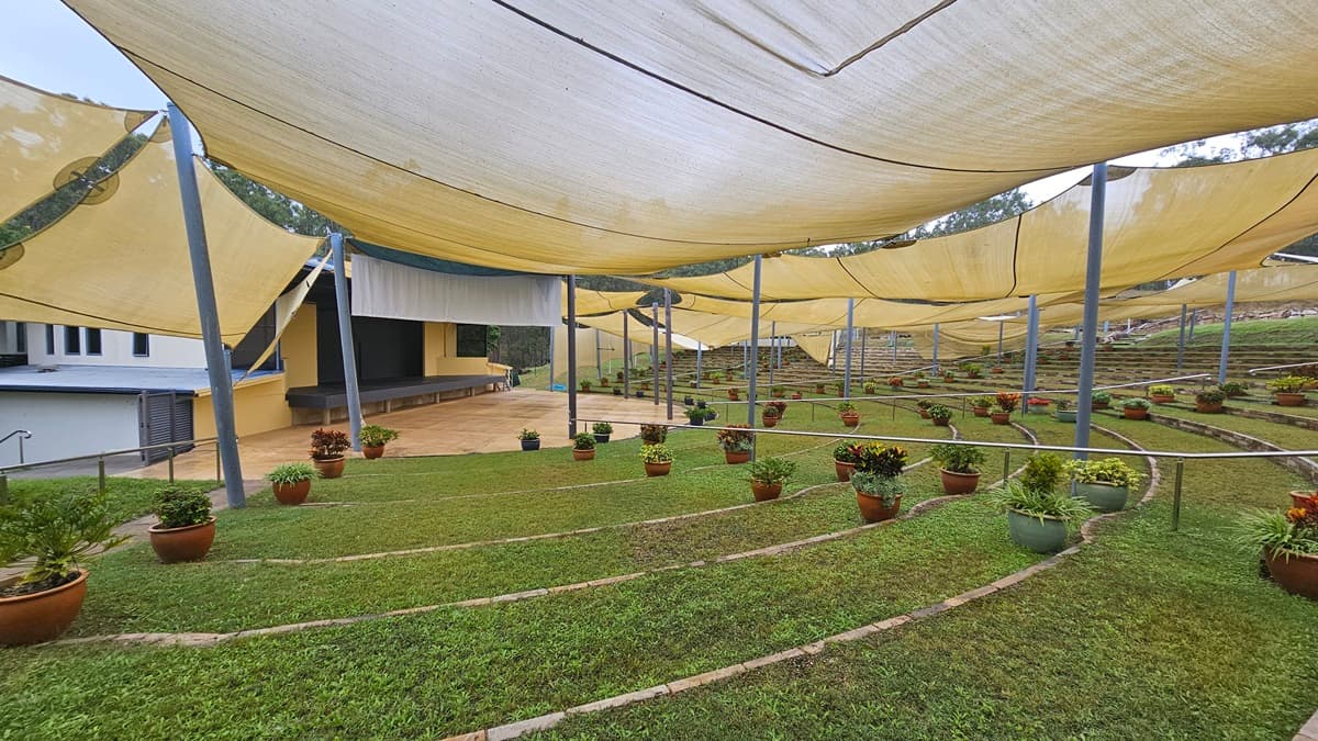 A terraced garden with neatly arranged potted plants on grassy steps. Overhead, large yellow shade cloths are stretched across metal poles, providing cover. In the background, a modern building with a sloped roof and a small stage area is visible. The ground is a mix of grass and paved paths, and the area is surrounded by a railing.
