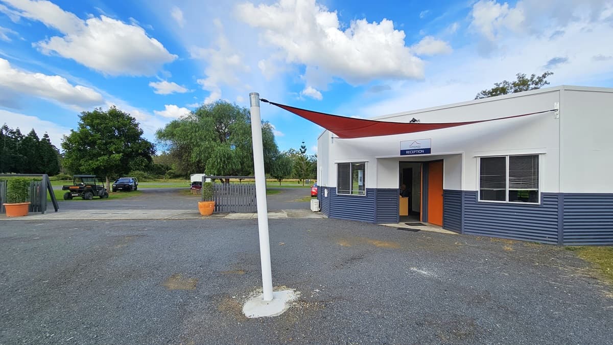 A small building with a sign reading "Reception" above the entrance is on the right. The building has a white and blue exterior with a red door. A red triangular shade sail is attached to a white pole in the foreground. On the left, there are trees and a few parked vehicles in the background. The ground is covered with gravel, and there are large potted plants near the entrance.