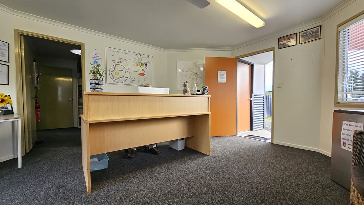 Reception area with a wooden desk in the centre. Behind the desk, a wall features a colourful map and a small plant. To the left, an open doorway leads to another room. On the right, an open door leads outside, with a view of a railing. The floor is carpeted in grey, and the ceiling has a fluorescent light fixture. A window with blinds is on the right wall, and framed certificates are visible on the left wall.