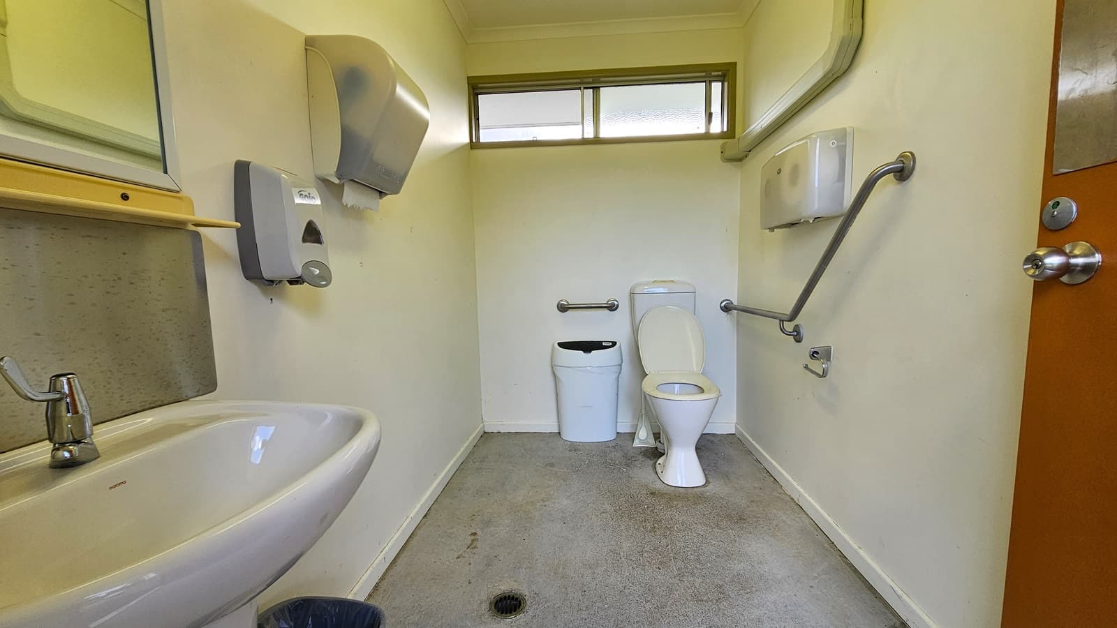 A small bathroom with a white toilet and a bin in the centre. On the left, there's a sink with a mirror above it, and a soap dispenser and hand dryer mounted on the wall. On the right, a grab rail is attached to the wall near the toilet, with a toilet paper holder beneath it. The floor is grey and slightly stained, and a narrow window is positioned high on the back wall. The door on the right has a metal handle.