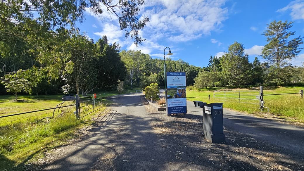 A rural road with a metal gate on the left and a grassy field on the right. In the centre, there's a signpost with a lamp above it, displaying information. Two letterboxes are positioned on the right side of the road. The background features dense trees and greenery under a partly cloudy sky. The ground is paved with some scattered leaves and debris.