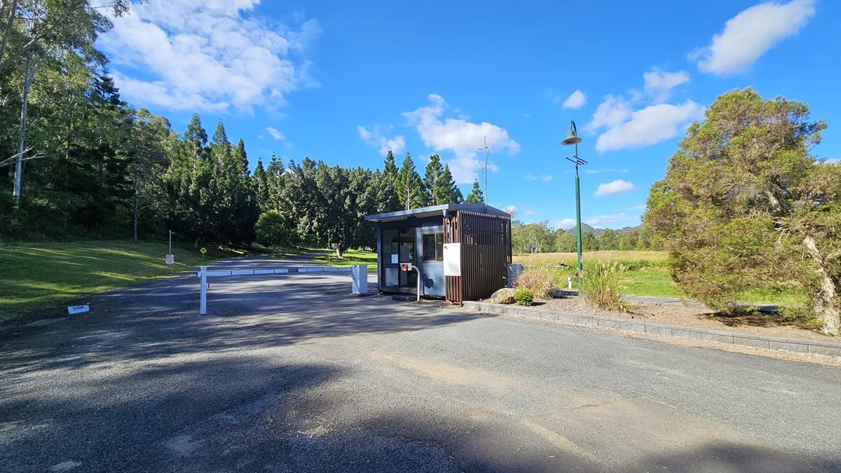 A small wooden booth is situated on the right side of a paved road, with a barrier gate in front. To the left, there is a grassy area with tall trees in the background. On the right, a lamp post stands near some bushes and a tree. The road is bordered by greenery, and the sky is clear with a few clouds.