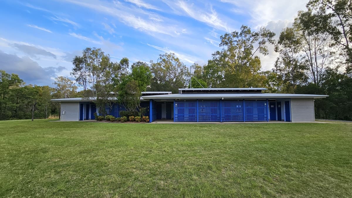 A modern, single-storey building with a flat roof and blue vertical slats on the facade. Solar panels are visible on the roof. The structure is surrounded by a well-maintained grassy lawn. Trees and shrubs are in the background, providing a natural setting. The building has a minimalist design with a focus on clean lines and functionality.