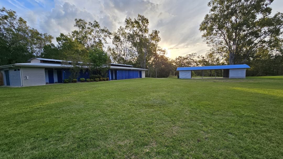 A grassy field with two structures. On the left, a long building with a flat roof and blue doors, surrounded by trees. On the right, a smaller open shelter with a blue roof. The background features tall trees and a setting sun.