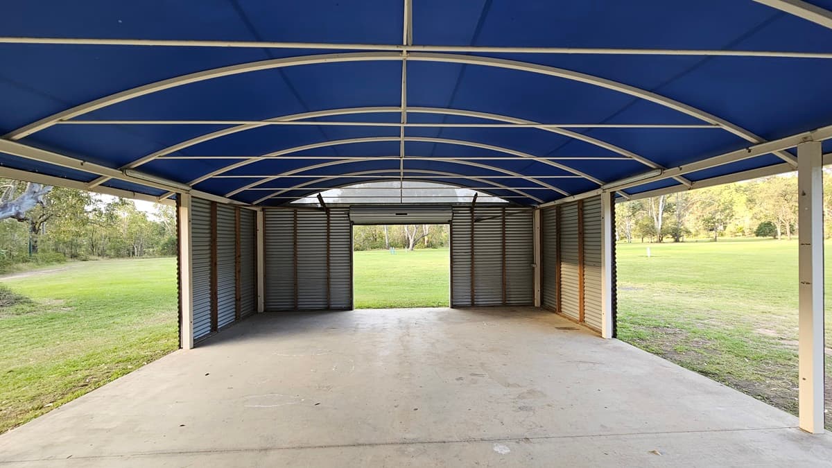 A large, open-sided shelter with a blue arched roof supported by a metal frame. The floor is concrete, and the walls are made of corrugated metal. The structure opens out to a grassy field with trees in the background. The ceiling features a series of curved beams. The left and right sides of the shelter are open, providing a view of the surrounding greenery.