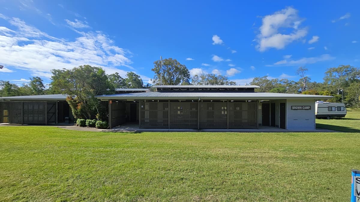A single-storey building with a flat roof and wooden slatted exterior. On the right, there's a sign that reads "Showers & Toilets." The building is surrounded by a well-maintained grassy area. In the background, there are trees and a caravan parked to the right. The sky is clear with a few clouds.