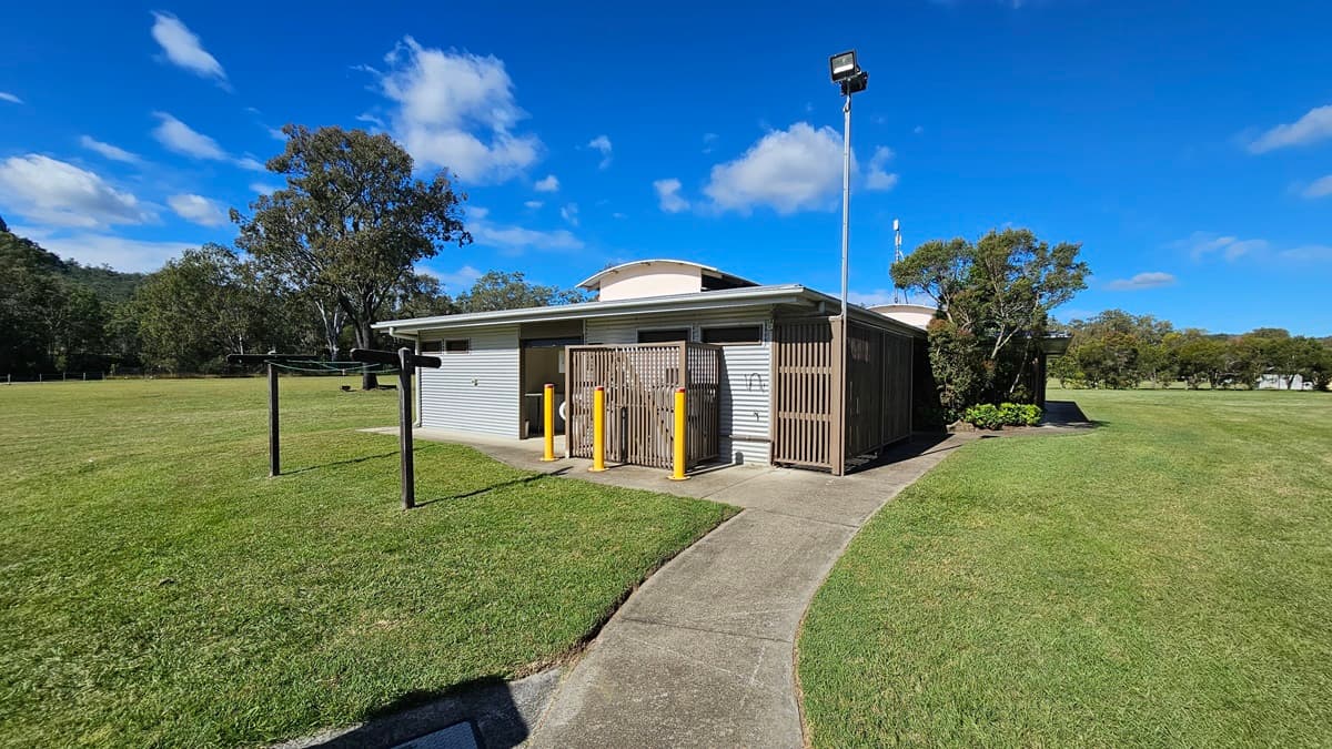 A small, single-storey building with a corrugated metal exterior and wooden slats on the front. Three yellow bollards stand in front of the entrance. A tall light pole is positioned to the right. The building is surrounded by a large grassy field with a few trees in the background. A concrete path leads up to the building, and there is a clothesline to the left. The sky is clear with a few clouds.