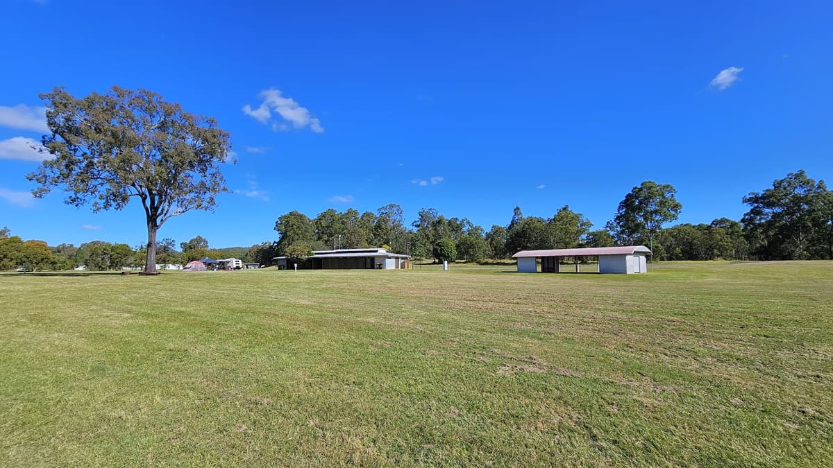 A large grassy field with a single tall tree on the left. In the middle distance, there are two small, simple buildings with flat roofs. The background is lined with dense trees. The sky is clear and blue with a few small clouds.