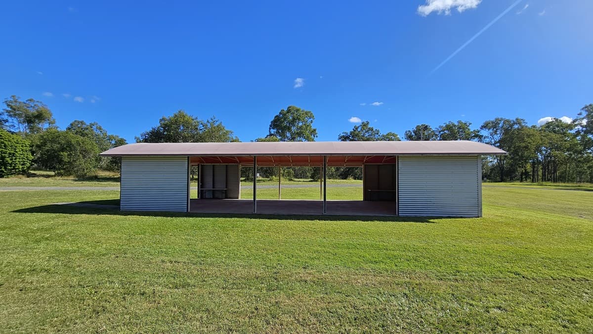 A small, rectangular structure with a corrugated metal exterior stands in the centre of a grassy field. The building has an open front with a roof supported by several vertical beams. Trees and shrubs are visible in the background, surrounding the field. The grass is neatly trimmed, and the sky is clear with a few clouds.