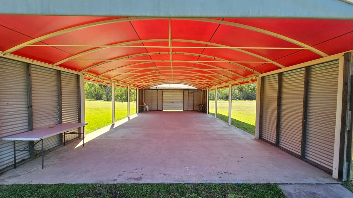 A long, open-sided structure with a red arched roof and a concrete floor. On the left, there is a folding table against a wall of closed metal shutters. The right side also features metal shutters. The background shows a grassy field and trees visible through the open sides. The ceiling is supported by metal beams.