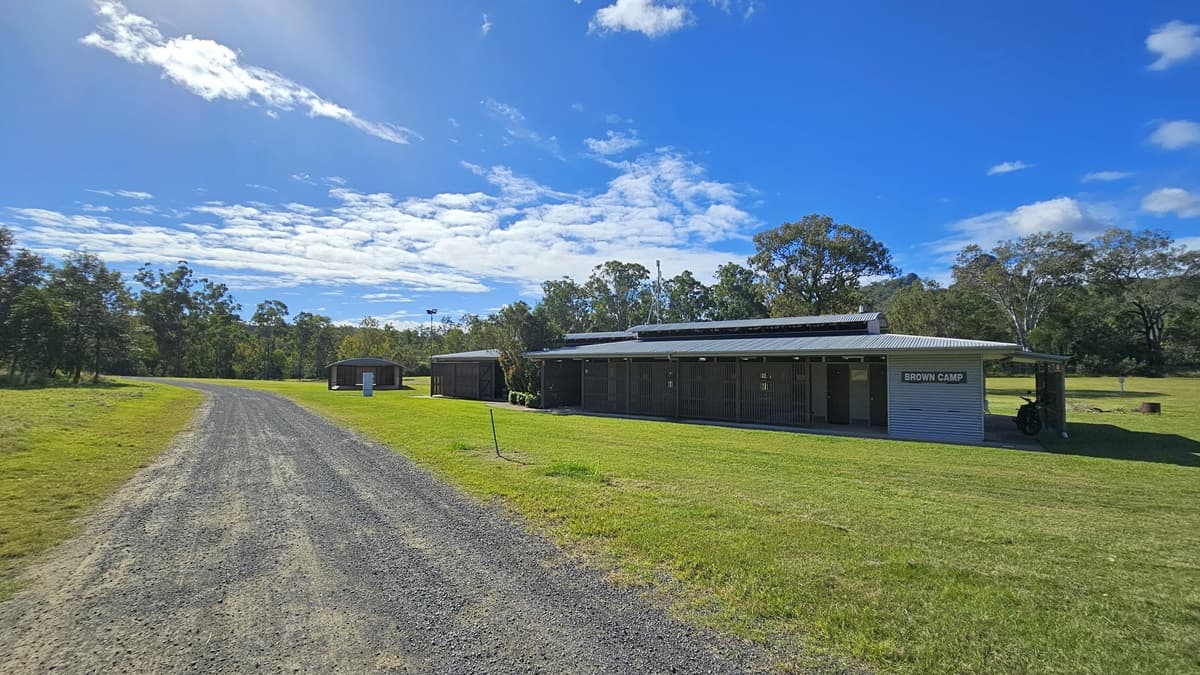 A gravel path runs from the foreground to the background on the left side, bordered by green grass. On the right, there is a long, single-storey building with a corrugated metal roof and a sign that reads "SHOWER BLOCK". Behind the building, there are trees and more greenery. The sky is mostly clear with some clouds.