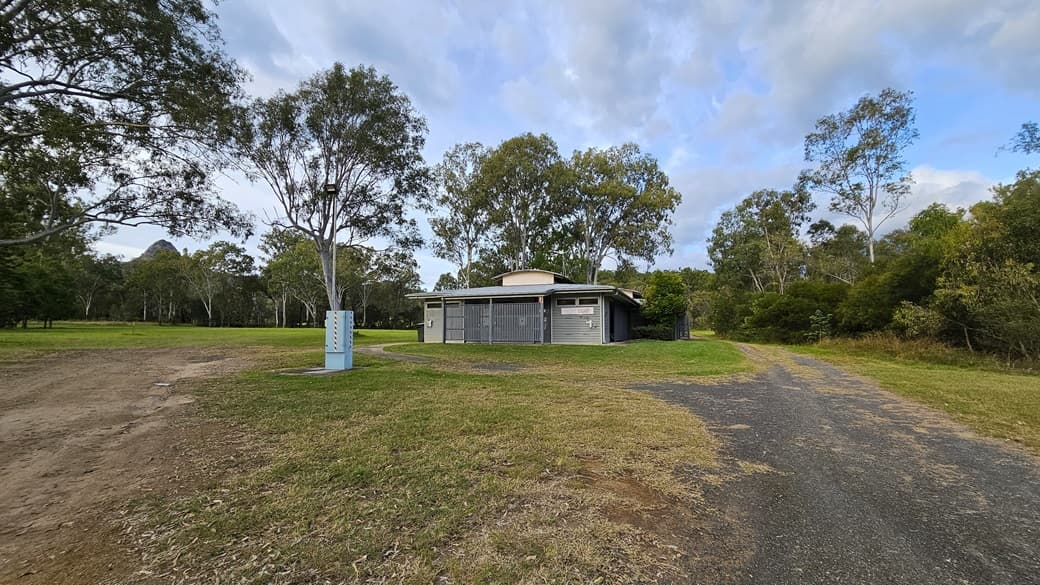A small, single-storey building with a flat roof is situated in the centre, surrounded by grass and trees. To the left, there is a dirt path and a utility box near a tree. On the right, a gravel path leads towards the building. The background is filled with tall trees and greenery. The sky is partly cloudy.