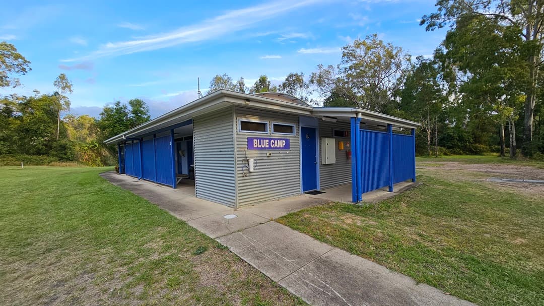 A small, single-storey building with a corrugated metal exterior and a sign reading "Blue Camp" on the front. The structure has blue panels and posts, with a concrete path leading to the entrance. Surrounding the building is a grassy area with trees in the background. The roof is flat, and the building appears to be in a natural, wooded setting.