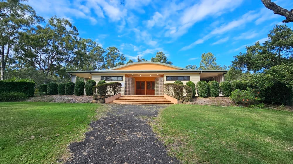 A single-storey house with a wide, sloping roof and a central entrance featuring wooden double doors. The house is surrounded by neatly trimmed hedges and bushes. A paved path leads up to the entrance, flanked by green lawns on either side. Tall trees are visible in the background, framing the house.