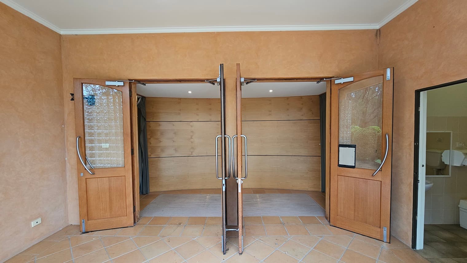 Two wooden doors with glass panels are open, leading into a room with a curved wall. The doors have metal handles. The floor is tiled in a light brown colour. On the right, there is a partially visible room with a sink and a toilet. The walls are a warm beige colour, and the ceiling is white with recessed lighting.