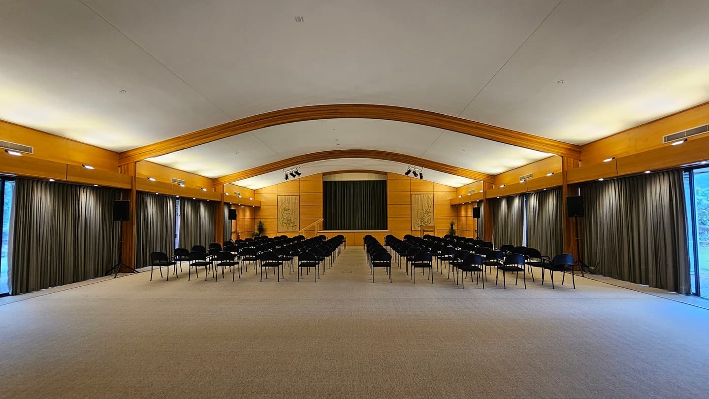 A spacious auditorium with a high, curved ceiling and wooden beams. Rows of black chairs are arranged facing a stage at the far end, which is flanked by two large decorative panels. The walls are lined with tall, dark curtains, and there are speakers mounted on the walls. The floor is carpeted in a light colour, and the room is well-lit with overhead lighting.