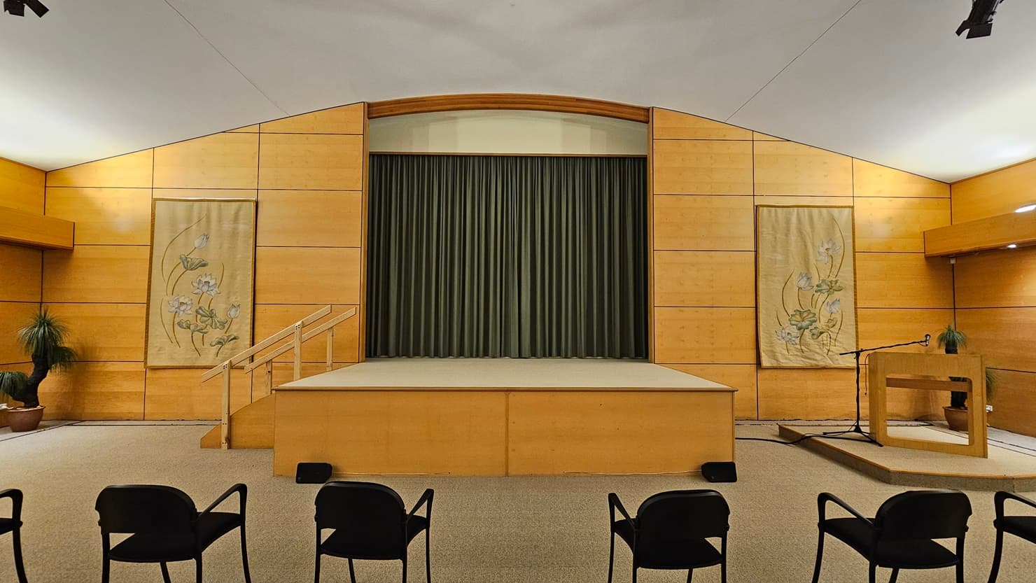 A room with a stage in the centre, featuring a green curtain backdrop. The walls are wooden with two decorative panels on either side of the stage, each displaying floral designs. On the right, there's a lectern with a microphone and a small plant. Several black chairs are positioned in the foreground. The ceiling is white, and the floor is carpeted. A potted plant is visible in the left corner.